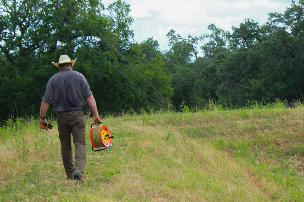 A man walks with a large measuring tape (known as an e line) with gloves in hand in a grassy field