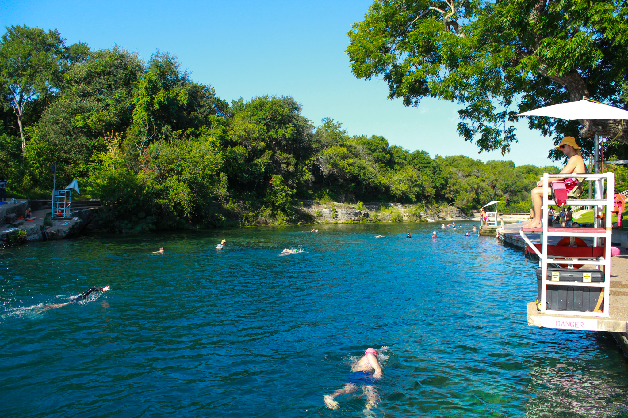 A view of Barton Springs pool will people swimming and a life guard on the side.
